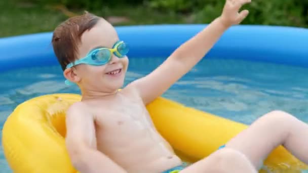 Happy baby boy in protective goggles floating on safety buoy at inflatable rubber swimming pool - Кадры, видео