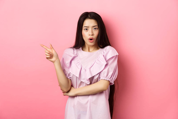 Scioccato asiatico giovanissima ragazza in abito indicando dito sinistro, ansimante meraviglia e guardare curioso a macchina fotografica, ponendo domanda su promo, in piedi su sfondo rosa - Foto, immagini