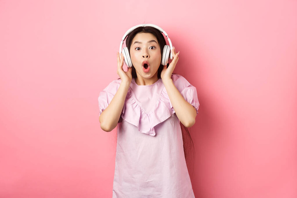Возбужденная азиатская девушка слышать удивительную песню, слушать музыку в наушниках и выглядеть изумленным, стоя на розовом фоне - Фото, изображение