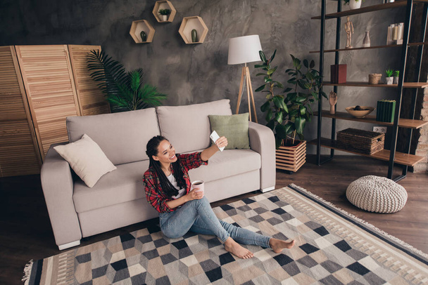 Portrait of attractive cheerful girl sitting on floor drinking caffeine resting taking selfie blogging at modern loft industrial home indoors - Photo, image