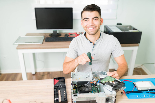 Boldog jóképű férfi mosolyog és szemkontaktust teremt miközben egy csavarhúzót tart, hogy megjavítson egy hibás CPU számítógépet a boltjában. - Fotó, kép