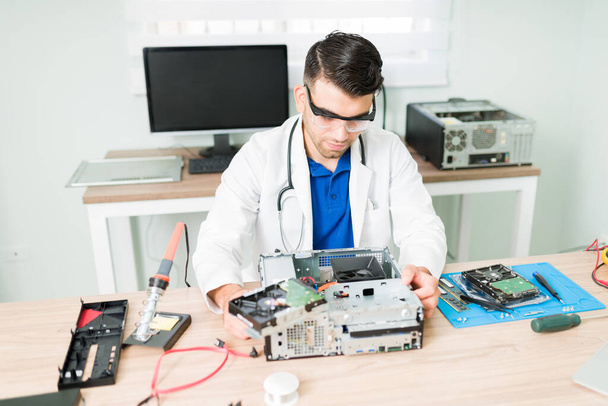 Un joven con una bata de laboratorio y gafas de protección arreglando una CPU rota. Técnico que trabaja como médico informático en el taller de reparación - Foto, imagen