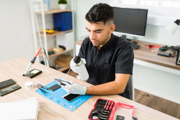 Jovem técnico do sexo masculino usando ar comprimido para limpar dentro de um smartphone sujo na oficina de reparação. Homem latino com luvas limpando um celular - Foto, Imagem