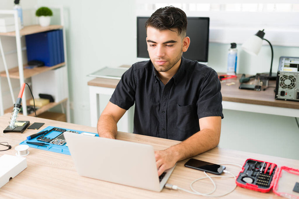 Schöner junger Mann, der die Software eines neuen Smartphones aktualisiert und auf seinem Laptop in der Werkstatt ein Datenbackup erstellt - Foto, Bild
