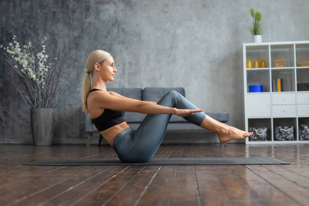 Young and sporty girl in sportswear is doing exercises in home interior. Fit and slender blond woman goes in for sports and fitness. Healthcare, fat burn and wellness. - 写真・画像