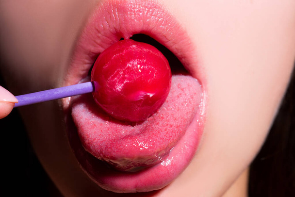 Girl with sexy mouth eating chupa chups close up. Woman lips sucking lollypop. Woman holding lollipop in mouth, close up. Red lips, sensual and sex shop concept. - 写真・画像