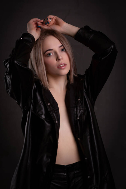 Gorgeous sexy blonde in a leather jacket dressed on a naked body, posing on a dark background with her hands up - Фото, зображення
