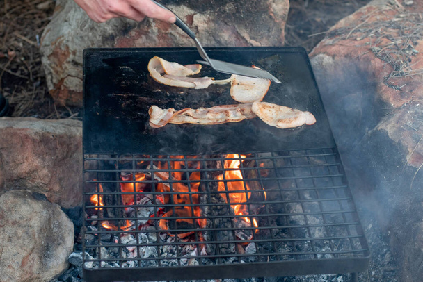 Breakfast camp cooking. Grilling crispy bacon on a cast iron plate over the camp fire. Camping lifesyle - Photo, Image