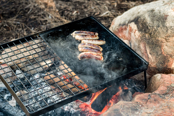 Camp cooking. Grilling sausages on a cast iron plate over the camp fire. Camping lifesyle - Photo, Image