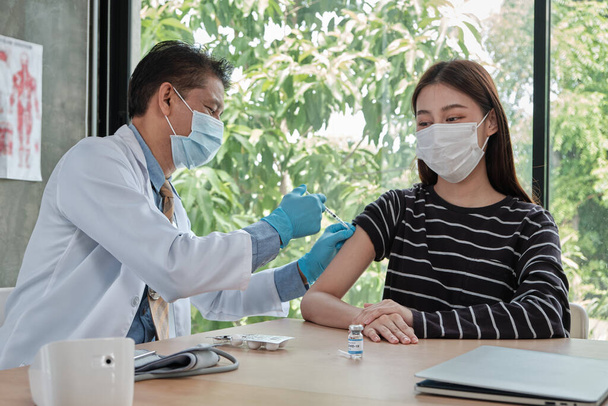 Mannelijke arts met gezichtsmasker vaccineren Aziatische vrouw, beschermen coronavirus (COVID19) in ziekenhuis kliniek. Injecties zijn een behandeling van ziekten, het voorkomen van infectie van de ziekte met behulp van gezonde medicijnen. - Foto, afbeelding