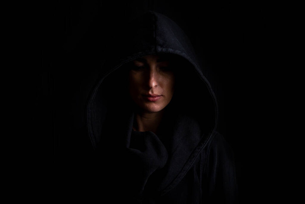 Νεαρή γυναίκα με μαύρη κουκούλα σε σκούρο μαύρο φόντο. - Φωτογραφία, εικόνα