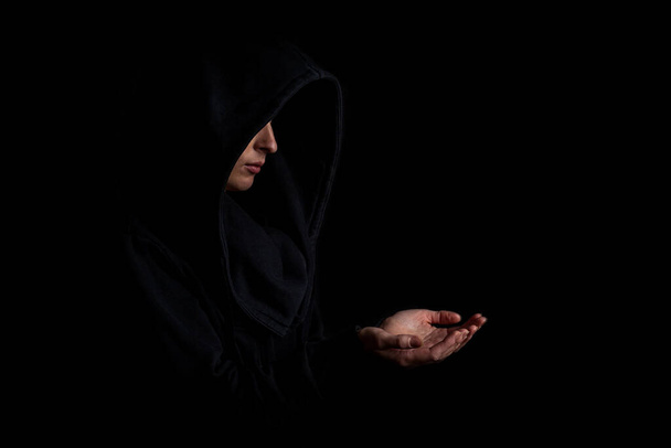 Νεαρή γυναίκα με μαύρη κουκούλα με απλωμένο χέρι σε σκούρο μαύρο φόντο. - Φωτογραφία, εικόνα
