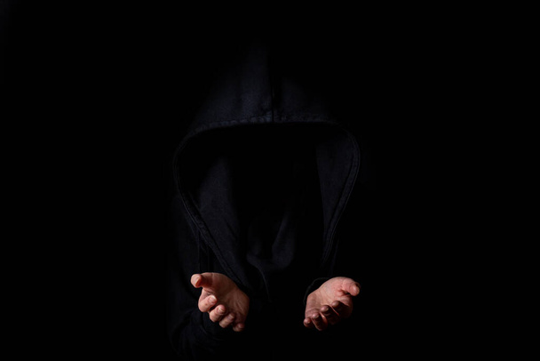 Junge Frau mit schwarzem Kapuzengesicht nicht sichtbar, Handflächen vor dunkelschwarzem Hintergrund ausgestreckt.  - Foto, Bild