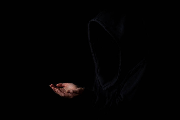 Mujer joven en la cara capucha negra no visible con las palmas extendidas sobre fondo negro oscuro. - Foto, Imagen