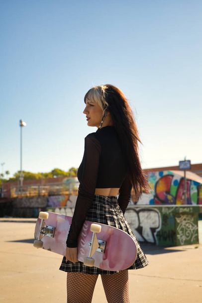 Young pretty girl with punk style and windblown hair holding a skateboard in her hand looking at infinity in profile at a skateboard park. Vertical position. - Photo, image