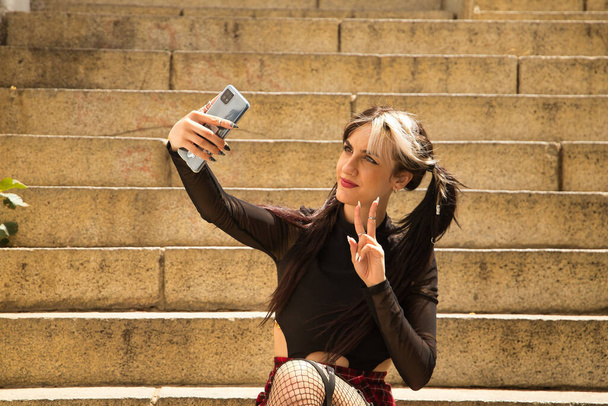 Young and beautiful girl with pigtails and punk style sitting on some stairs taking a selfie with her mobile phone while making the victory sign. - Photo, Image