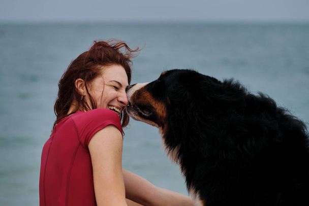 Caucasian pretty red haired woman and Bernese Mountain Dog embrace against background of blue sea and enjoy each other. Walking and relaxing with large dog on warm summer morning. - Foto, Bild