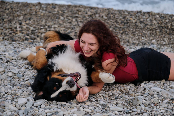 Caucasian pretty red haired woman scratches belly of Bernese Mountain Dog and laughs merrily and sincerely. Joyful happy walk and rest with large dog on warm summer morning. - Фото, зображення