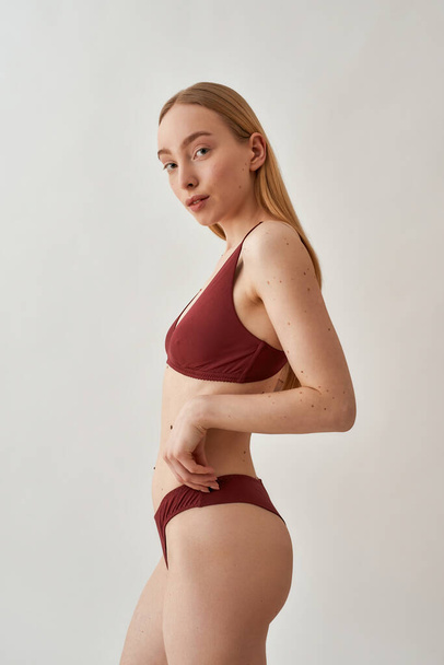 Side view of young caucasian woman with slim body wearing burgundy underwear looking at camera, standing isolated over light gray background - Fotografie, Obrázek