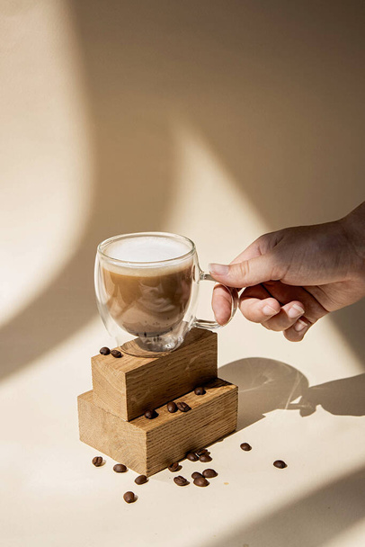 Kaffee in einer Glasschale auf beigem Hintergrund. Die Hand hält einen Becher. Minimalistische stilvolle Fotografie bei Tageslicht mit Schatten und Licht. - Foto, Bild