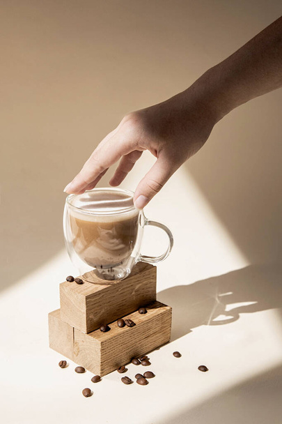 Café em uma xícara de vidro em um fundo bege. A mão segura um copo. Fotografia minimalista e elegante à luz do dia com sombras e luz. - Foto, Imagem