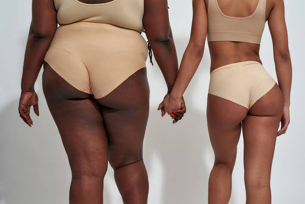 Oříznutý záběr dvou afrických amerických žen ve spodním prádle s různou velikostí těla drží se za ruce, stojí izolované nad šedým pozadím - Fotografie, Obrázek