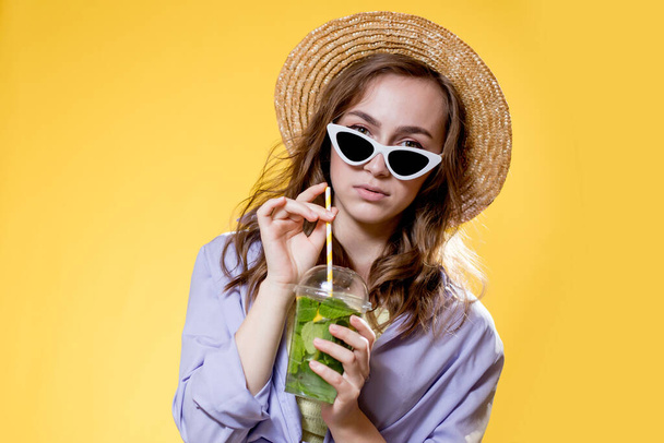 Girl in summer outfit wearing white trendy sunglasses and enjoying while drinking a Mojito cocktail. Posing on the yellow background. - Zdjęcie, obraz