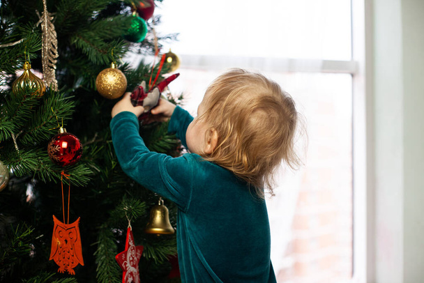 little baby hangs a toy on a christmas tree. New year and christmas at home. Winter Holidays. Decorate the Christmas tree - Fotó, kép