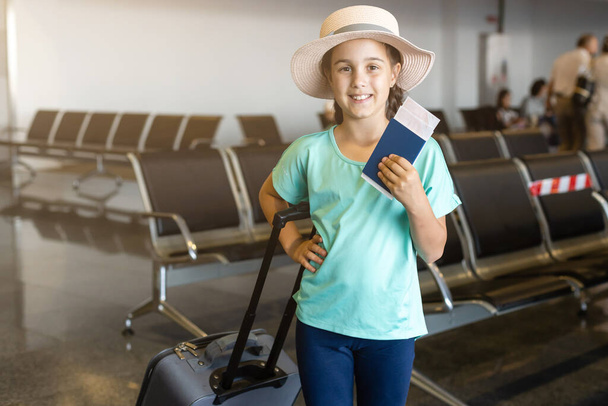 τουρισμός, διακοπές, παιδική ηλικία και μεταφορά έννοια - χαμογελαστό κοριτσάκι με ταξιδιωτική τσάντα, εισιτήριο και διαβατήριο πάνω από το αεροδρόμιο φόντο. - Φωτογραφία, εικόνα