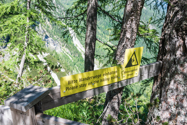 登山道にはドイツ語と英語で黒い文字の木に照らされた黄色の警告記号が言っています。Bitte Wanderwege einhalten, abstrzgefahr,ハイキングコースに滞在してください,落下のリスク. - 写真・画像