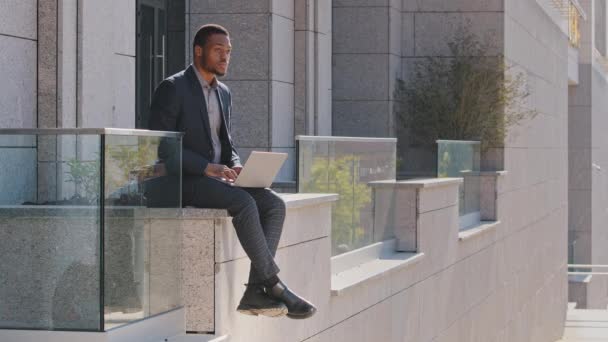 Pensativo hombre de negocios de raza mixta hombre negro millennial con traje sentado al aire libre escribiendo en el ordenador portátil, preocupado por el problema, chico pensando en tomar decisiones difíciles mirando hacia otro lado, la estrategia de planificación - Metraje, vídeo