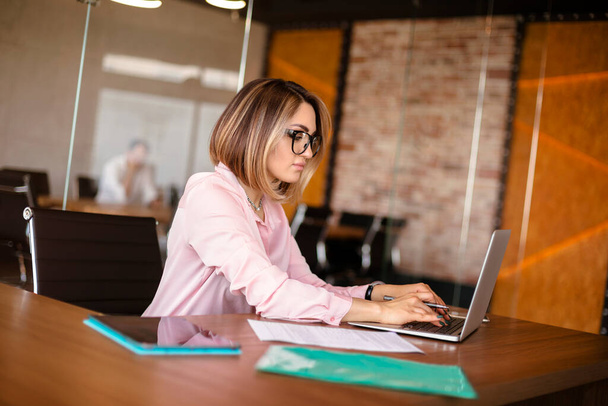 Молодая женщина-менеджер в очках, печатающая на нетбуке, сидя за столом и работая в современном офисе - Фото, изображение