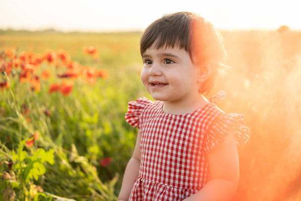 close up portrait beautiful happy baby girl in red dress smiling on field of poppies at summer sunset - Valokuva, kuva