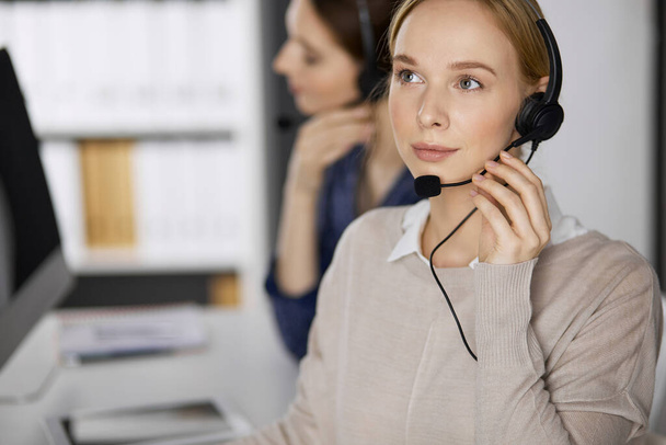 Friendly businesswoman talking by headset while sitting in office. Call center and diverse people group in business - Фото, изображение
