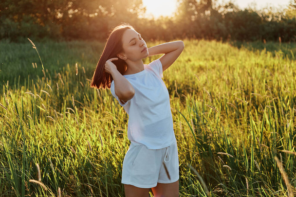 Outdoor shot of attractive dark haired female wearing white t shirt and short looking away, raising arms, posing in green meadow, enjoying beautiful sunset and nature. - Foto, immagini