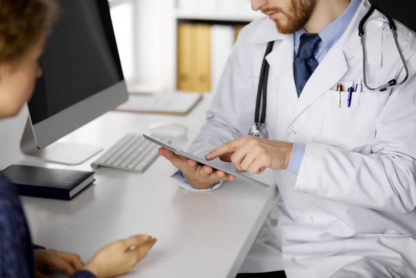 Unknown bearded doctor and patient woman discussing current health examination while sitting and using tablet computer, close-up. Medicine concept - Fotoğraf, Görsel