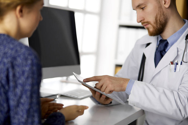 Unknown bearded doctor and patient woman discussing current health examination while sitting and using tablet computer, close-up. Medicine concept - Foto, afbeelding