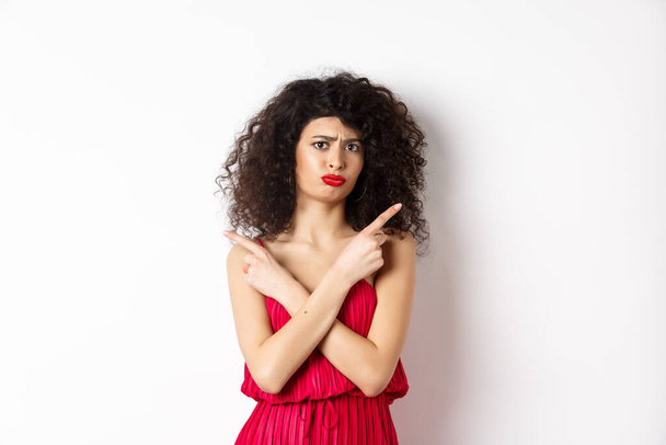 Doubtful and sad young woman in red dress, pointing fingers sideways and sulking, cant decide between products, need help with choice, standing over white background - 写真・画像