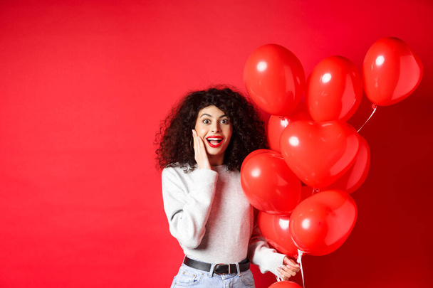 Valentinstag. Überrascht lockige Mädchen mit Herz Luftballons und Blick in die Kamera, erhalten Überraschungsgeschenk auf Datum von Liebhaber, steht auf rotem Hintergrund - Foto, Bild