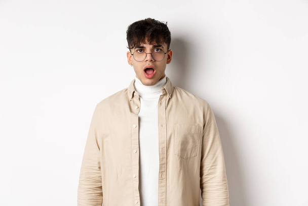 Portrait of shocked young man in glasses drop jaw, open mouth and stare amazed at camera, looking at something impressive, standing on white background - Фото, изображение
