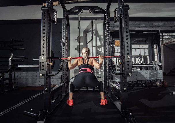 Μια γυμνάστρια κάθεται με μια καμπάνα στους ώμους της στο σκοτεινό γυμναστήριο. Τρόπος ζωής, bodybuilding, άρση βαρών και καλή φυσική κατάσταση - Φωτογραφία, εικόνα