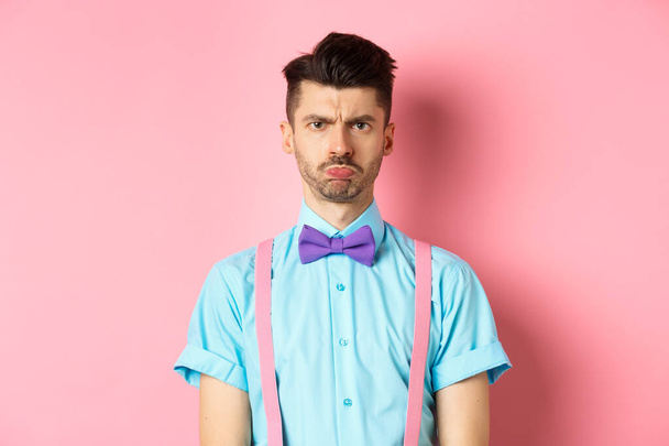 Θλιβερός και προσβεβλημένος νεαρός άνδρας, συνοφρυωμένος και σκυθρωπός χείλη, κοιτάζοντας θυμωμένος με κάποιον, στέκεται απογοητευμένος σε ροζ φόντο με παπιγιόν - Φωτογραφία, εικόνα
