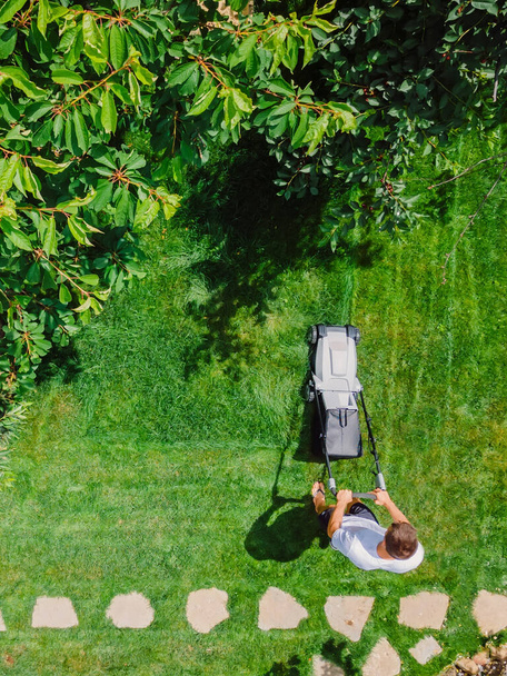 Kaukasische tuinman maaien achtertuin gras met behulp van draadloze elektrische grasmaaier. Luchtfoto 's. Tuinieren en landschapsarchitectuur. - Foto, afbeelding