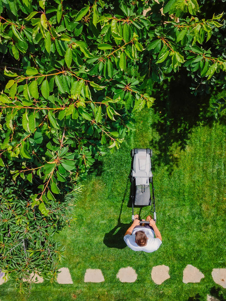 Καυκάσιος κηπουρός κουρεύει γρασίδι κήπου χρησιμοποιώντας ηλεκτρικό χλοοκοπτικό μπαταρίας. Αεροφωτογραφία. Βιομηχανία κηπουρικής και εξωραϊσμού. - Φωτογραφία, εικόνα