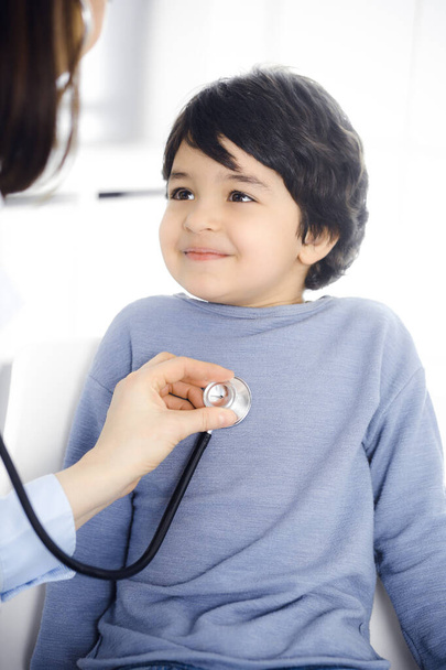 Doctor-woman examining a child patient by stethoscope. Cute arab boy at physician appointment. Medicine concept - Foto, Imagen