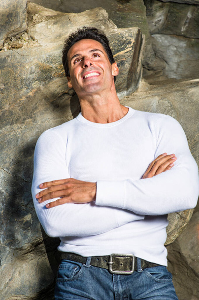 Habillé d'un pull blanc, croisant les bras, un homme d'âge moyen est debout près des rochers, regardant joyeusement vers le haut - Photo, image