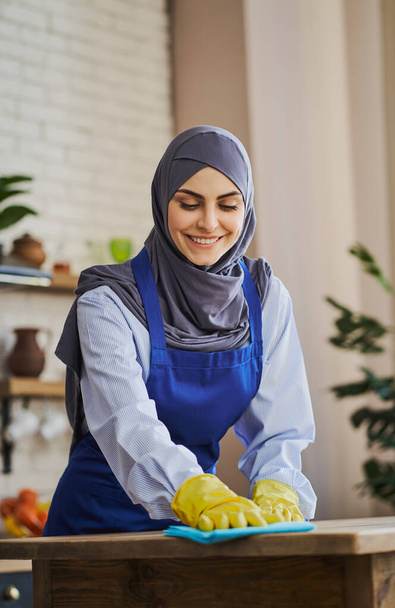 Pretty Arabian woman in apron and gloves cleaning a table in the kitchen - Fotó, kép
