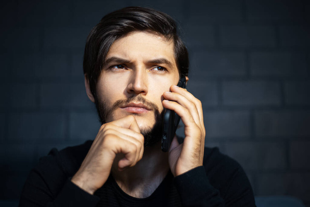 Thoughtful young man talking on smartphone on background of black brick wall. - Valokuva, kuva