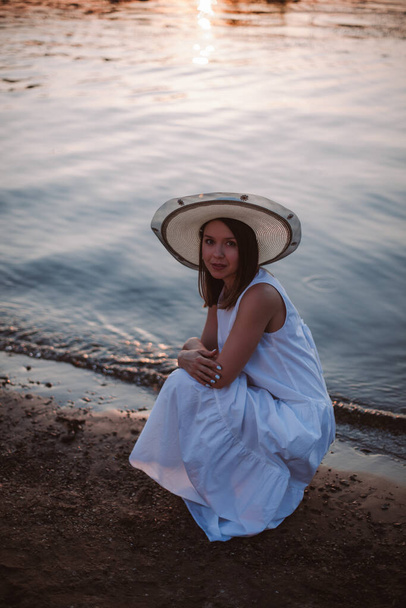 a happy Caucasian young woman in a white sundress and hat is squatting and hugging her shoulders on the beach at sunset on a summer evening - Fotografie, Obrázek