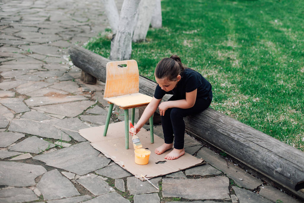 portrait of a full-length child painting a chair for reuse and sitting on a log on a stone path by the lawn on a summer day - Fotoğraf, Görsel